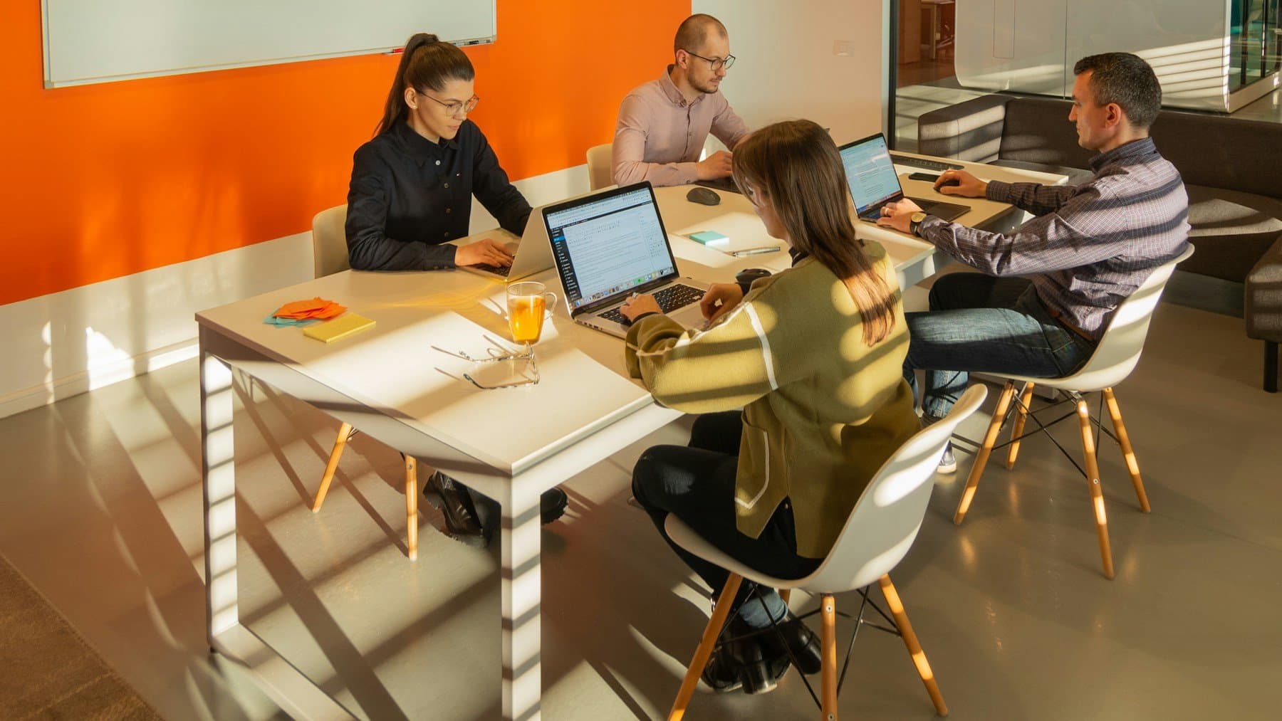 Four colleagues working on laptops around a shared table in a bright meeting room with an orange wall